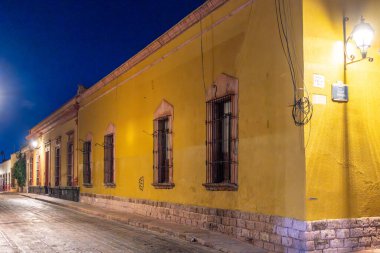 A yellow building with a dark street in front of it. The street is lit up by street lights. Historic center of Queretaro, colonial architecture, decorations for the celebration of Mexico's Independence Day.