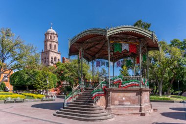 A small gazebo with a large tower in the background. The gazebo is decorated with colorful flags. Historic center of Queretaro, colonial architecture, decorations for the celebration of Mexico's Independence Day.