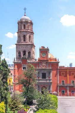 A large red building with a clock tower and a spire. The building is surrounded by trees. Historic center of Queretaro, colonial architecture, decorations for the celebration of Mexico's Independence Day.