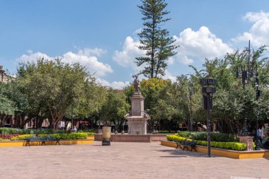A large statue of a man stands in front of a fountain in a park. The statue is surrounded by trees. Historic center of Queretaro, colonial architecture, decorations for the celebration of Mexico's Independence Day.