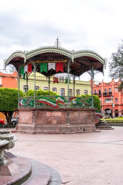 Arkasında beyaz bir bina olan küçük bir kasaba meydanı. Binanın üzerinde bir bayrak var. Queretaro 'nun tarihi merkezi, sömürge mimarisi, Meksika' nın Bağımsızlık Günü kutlamaları için dekorasyonlar.