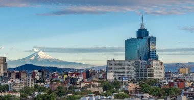 A city skyline with a large building in the background and a mountain in the distance. The sky is clear and blue. Panoramic view of Mexico City, snow-covered volcanoes Popocatepetl and Iztaccihuatl