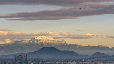 Uzaktan görülebilen bir dağ sırası ve üzerinde uçan bir uçak var. Gökyüzü bulutlu ve güneş batıyor. Mexico City 'nin panoramik manzarası, karla kaplı volkanlar Popocatepetl ve Iztaccihuatl!
