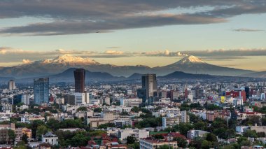 A city with a mountain range in the background. The city is full of tall buildings and the sky is cloudy. Panoramic view of Mexico City, snow-covered volcanoes Popocatepetl and Iztaccihuatl