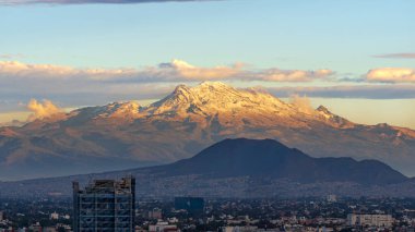 A mountain range is visible in the distance behind a city. The sky is cloudy and the sun is setting. Panoramic view of Mexico City, snow-covered volcanoes Popocatepetl and Iztaccihuatl