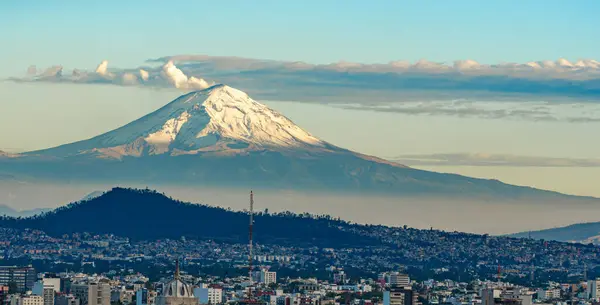 A mountain with a snow covered peak is in the background of a city. The city is bustling with activity. Panoramic view of Mexico City, snow-covered volcanoes Popocatepetl and Iztaccihuatl