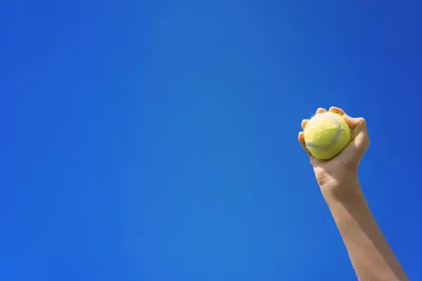 Yellow tennis ball in a girl's hand against a blue sky background. Indispensable sports attribute for playing tennis. Winning gesture of a tennis player. Sports gear for tennis