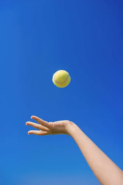 A hand tosses a tennis ball into the sky. A hand tosses a tennis ball for a serve against a blue sky. Sports equipment for tennis