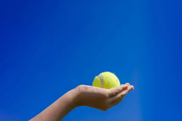 Mavi arka planda bir kızın elinde sarı tenis topu. Tenis oynamak için zorunlu spor ekipmanı. El, maç başlamadan önce bir tenis topu tutuyor. Tenis için spor malzemeleri.
