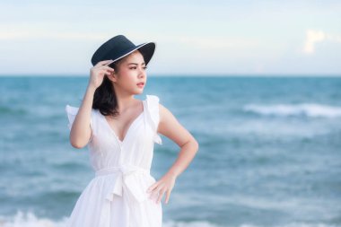 Beyaz elbiseli ve siyah şapkalı genç Asyalı kadın turist sahilde rahatlayıp, denizin ve yaz tatilinin tadını çıkarıyor.,