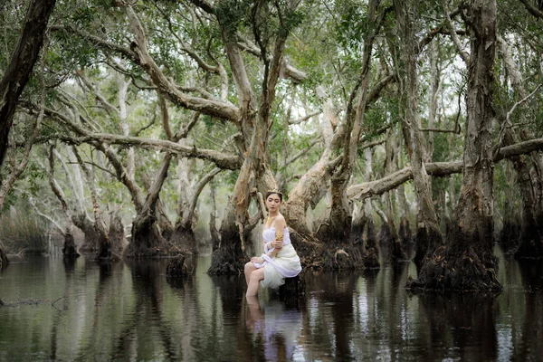 美丽动人的泰籍亚洲女人穿着白色的当地传统服装 名字叫Nakee 坐在湖边的树桩上 在植物园里 Rayong Thailand Nakee Portait模型在公园里的概念 — 图库照片