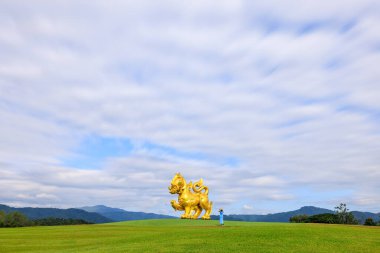  young woman tourist standing using smartphone taking Singha statue and clouds sky background, at Singha Park, Chiangrai, Thailand. Gold Lion statue in Boonrawd farm.