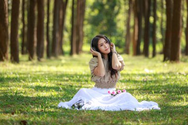 beautiful potrait asian woman siting and listening to headphone music in a pine forest and retro camera with rose flowers in frame, lifestyle and freedom in vacation concept,