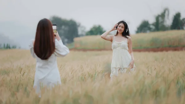 在金黄色的大麦中散步时 两个身穿白色连衣裙 拿着自拍和耳机无线的年轻漂亮的女人高兴地微笑着 — 图库照片