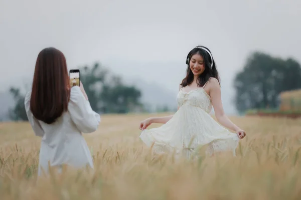 在金黄色的大麦中散步时 两个身穿白色连衣裙 拿着自拍和耳机无线的年轻漂亮的女人高兴地微笑着 — 图库照片