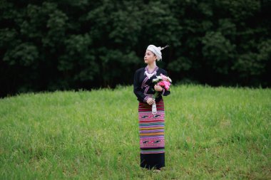 Asyalı genç bir kadının doğal ışık portresi yeşil çimlerin üzerinde duruyor geleneksel Tayland kültürü giysisi tarzı giyiniyor ve ellerinde çiçekler, orman arka planında.