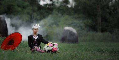 Asyalı genç bir kadının doğal ışık portresi yeşil çimenlerde oturmuş geleneksel Tayland kültürü elbisesi, çiçek sepeti ve doğal bahçedeki kırmızı şemsiyesiyle orman arka planında.