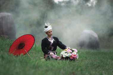 Asyalı genç bir kadının doğal ışık portresi yeşil çimenlerde oturmuş geleneksel Tayland kültürü elbisesi, çiçek sepeti ve doğal bahçedeki kırmızı şemsiyesiyle orman arka planında.