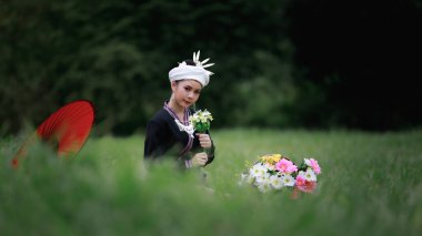 Asyalı genç bir kadının doğal ışık portresi yeşil çimenlerde oturmuş geleneksel Tayland kültürü elbisesi, çiçek sepeti ve doğal bahçedeki kırmızı şemsiyesiyle orman arka planında.