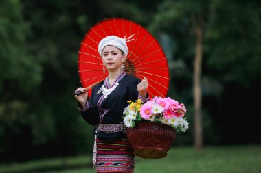 Asyalı genç bir kadının portresi yeşil çimlerin üzerinde geleneksel Tayland kültürü elbisesi, çiçek sepeti ve doğal bahçedeki kırmızı şemsiyesiyle orman arka planında duruyor.