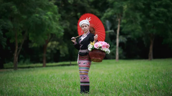 亚洲年轻女子的画像 站在绿草上 身穿传统的泰族文化长裙 穿着花篮和红色的雨伞 在自然的花园 森林的背景 — 图库照片