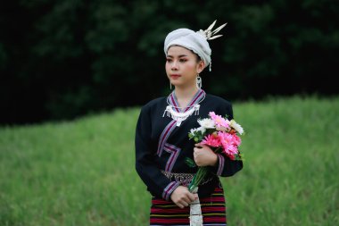 Asyalı genç bir kadının doğal ışık portresi yeşil çimlerin üzerinde duruyor geleneksel Tayland kültürü giysisi tarzı giyiniyor ve ellerinde çiçekler, orman arka planında.