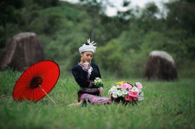 Asyalı genç bir kadının doğal ışık portresi yeşil çimenlerde oturmuş geleneksel Tayland kültürü elbisesi, çiçek sepeti ve doğal bahçedeki kırmızı şemsiyesiyle orman arka planında.