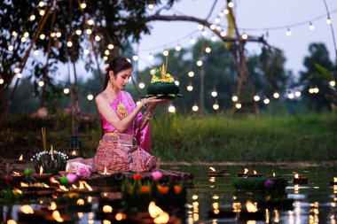 Loi Krathong Festivali, Taylandlı bir kadın nehir kenarında bir salda oturmuş krathong tutuyor, geleneksel Tayland kostümlü Asyalı kadınlar Loi Krathong Günü, gelenekler ve Tayland kültüründe yüzmek üzere kratonglar getiriyor.