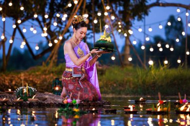 Loi Krathong Festivali, Taylandlı bir kadın nehir kenarında bir salda oturmuş krathong tutuyor, geleneksel Tayland kostümlü Asyalı kadınlar Loi Krathong Günü, gelenekler ve Tayland kültüründe yüzmek üzere kratonglar getiriyor.