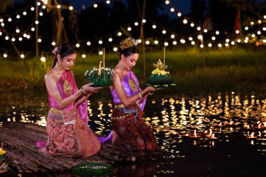 Loi Krathong Festivali, nehir kenarında bir salda oturan iki Taylandlı kadın, geleneksel Tayland kostümlü Asyalı kadınlar, Tayland gelenek ve kültüründe, Loi Krathong Günü 'nde yüzmek üzere kratonglar getiriyor.,
