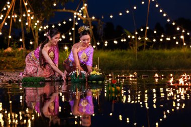 Loi Krathong Festivali, nehir kenarında bir salda oturan iki Taylandlı kadın, geleneksel Tayland kostümlü Asyalı kadınlar, Tayland gelenek ve kültüründe, Loi Krathong Günü 'nde yüzmek üzere kratonglar getiriyor.,