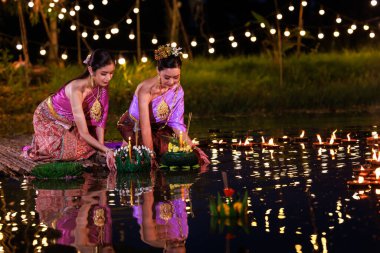 Loi Krathong Festivali, nehir kenarında bir salda oturan iki Taylandlı kadın, geleneksel Tayland kostümlü Asyalı kadınlar, Tayland gelenek ve kültüründe, Loi Krathong Günü 'nde yüzmek üzere kratonglar getiriyor.,