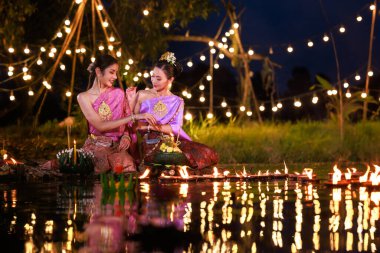 Loi Krathong Festivali, nehir kenarında salda oturan iki Taylandlı kadın, geleneksel Tayland kostümlü Asyalı kadınlar, Loi Krathong Günü 'nde yüzmek üzere kramponlar getiriyor, Tayland gelenekleri ve kültürü.,