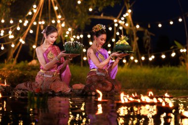 Nehir kenarında bir salda oturan iki Taylandlı kadın, geleneksel Tayland kostümlü Asyalı kadınlar, Loi Krathong Günü 'nde, gelenek ve Tayland kültüründe yüzmek üzere krathonglar getirirler.