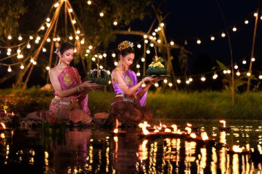 Loi Krathong Festivali, nehir kenarında bir salda oturan iki Taylandlı kadın, geleneksel Tayland kostümlü Asyalı kadınlar, Tayland gelenek ve kültüründe, Loi Krathong Günü 'nde yüzmek üzere kratonglar getiriyor.,