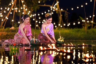 Loi Krathong Festivali, nehir kenarında bir salda oturan iki Taylandlı kadın, geleneksel Tayland kostümlü Asyalı kadınlar, Tayland gelenek ve kültüründe, Loi Krathong Günü 'nde yüzmek üzere kratonglar getiriyor.,