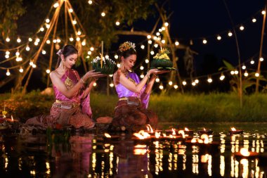 Loi Krathong Festivali, nehir kenarında bir salda oturan iki Taylandlı kadın, geleneksel Tayland kostümlü Asyalı kadınlar, Tayland gelenek ve kültüründe, Loi Krathong Günü 'nde yüzmek üzere kratonglar getiriyor.,