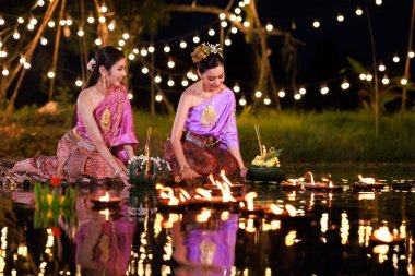 Loi Krathong Festivali, nehir kenarında bir salda oturan iki Taylandlı kadın, geleneksel Tayland kostümlü Asyalı kadınlar, Tayland gelenek ve kültüründe, Loi Krathong Günü 'nde yüzmek üzere kratonglar getiriyor.,