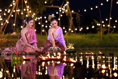 Nehir kenarında bir salda oturan iki Taylandlı kadın, geleneksel Tayland kostümlü Asyalı kadınlar, Loi Krathong Günü 'nde, gelenek ve Tayland kültüründe yüzmek üzere krathonglar getirirler.