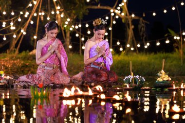 İbadet eden ve dua eden iki Taylandlı kadın nehir kenarında bir salda oturup dua ediyorlar. Geleneksel Tayland kostümlü Asyalı kadınlar, Loi Krathong Günü 'nde yüzmek üzere kramponlar getiriyorlar. Gelenekler ve Tayland kültürü.