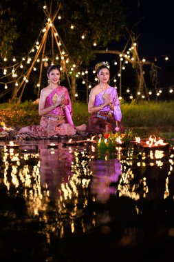 Loi Krathong Festivali, nehir kenarında bir salda oturan iki Taylandlı kadın, geleneksel Tayland kostümlü Asyalı kadınlar, Tayland gelenek ve kültüründe, Loi Krathong Günü 'nde yüzmek üzere kratonglar getiriyor.,