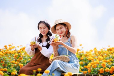 İki kadın çiçek bahçesinde elinde mandalina ya da portakal meyvesi tutarak kameraya bakıyor.