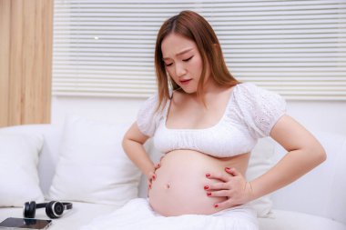 Asyalı hamile bir kadın yatak odasında oturmuş karnına dokunuyor karnında ağrı hissediyor.