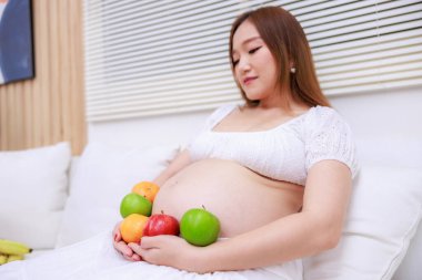 Kanepede oturan Asyalı hamile kadın, elinde elma, turuncu, seçici bir odak noktası var..