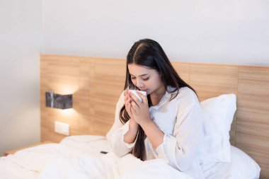 Genç Asyalı kadın sabah vakti yatakta oturup kahve içiyor., 