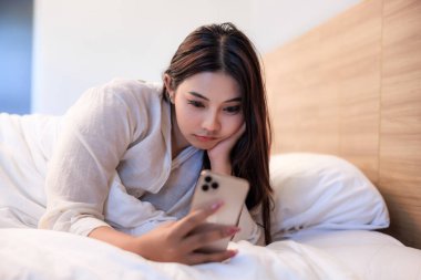 Asyalı bir kadın yatak odasındaki bir dairede yatağında telefonunda bir mesaj görünce rahatsız ve stresli olur..