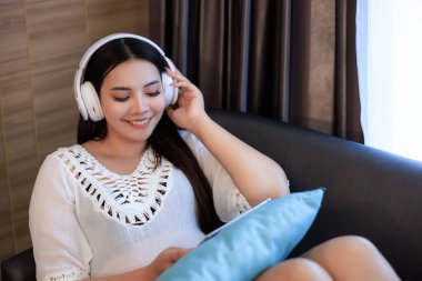 Genç Asyalı kadın kulaklık takıyor evde dinleniyor ve kanepede müzik dinliyor.