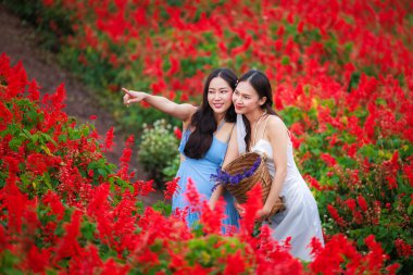 Kızıl çiçek bahçesinde poz veren ve elinde sepet tutan iki genç kadının portresi.. 