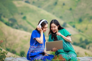 Kabile kıyafetleri giymiş iki genç Asyalı çayırda dizüstü bilgisayarını kullanarak mutlu mesut alışveriş yapıyor. Doğa tepesi geçmişleri.