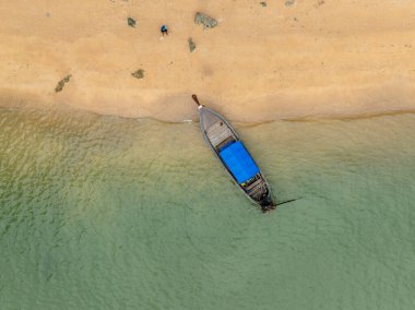 Zümrüt yeşili su ve altın kumu olan bir sahile yanaşmış uzun kuyruklu bir teknenin havadan görüntüsü. Kıyıda dalgaların izleri ve uzak bir insan var. Bu Tayland denizlerinin ve kıyı yaşamının güzelliğini yansıtır..